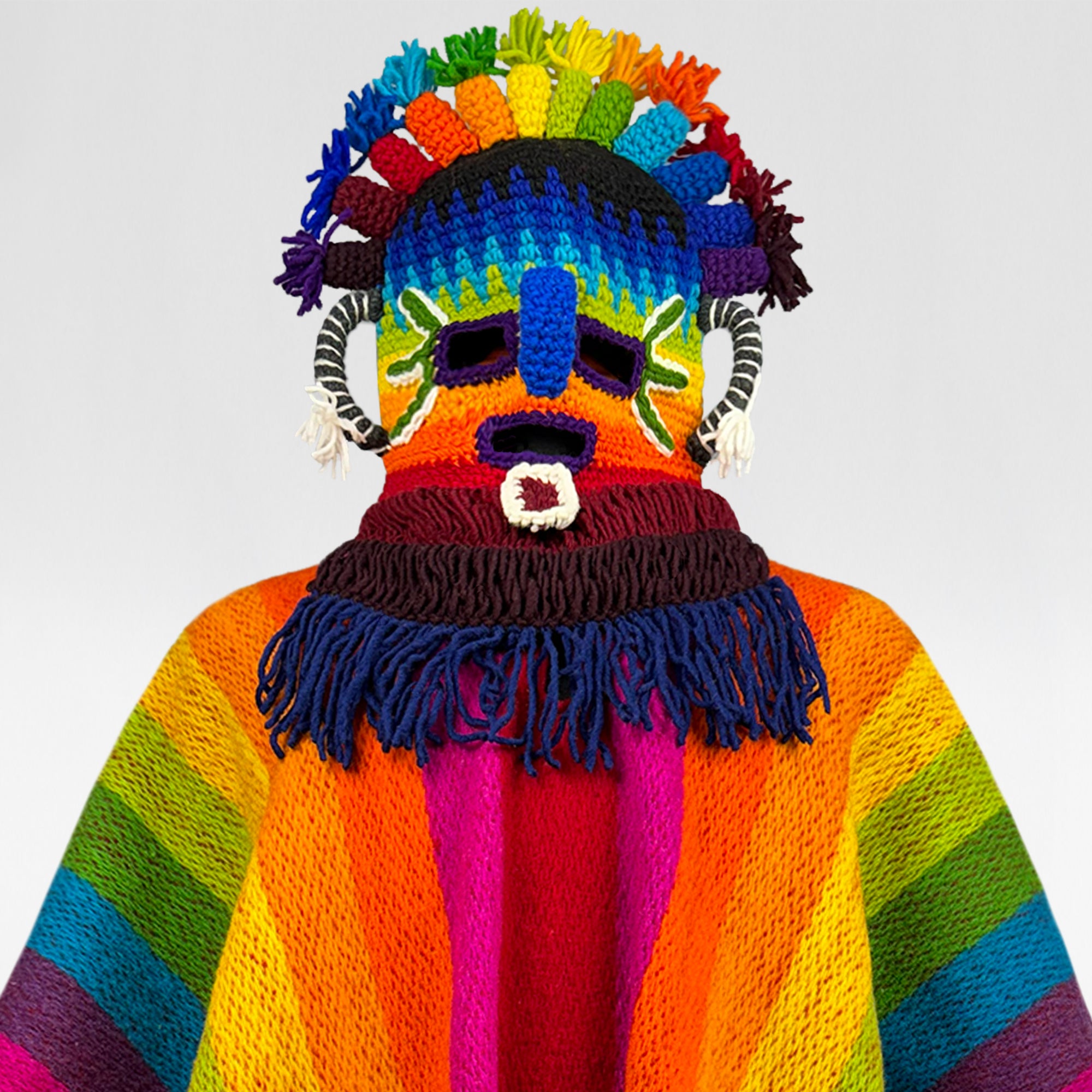 A colorful knitted mask covers a person's head and shoulders, featuring vibrant stripes and patterns in rainbow hues. The mask includes facial features like eyes and nose, framed by fringed yarn.