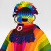 A knitted mask with vibrant rainbow colors covers a figure's head, adorned with fringe and spiral designs, set against a plain white background.