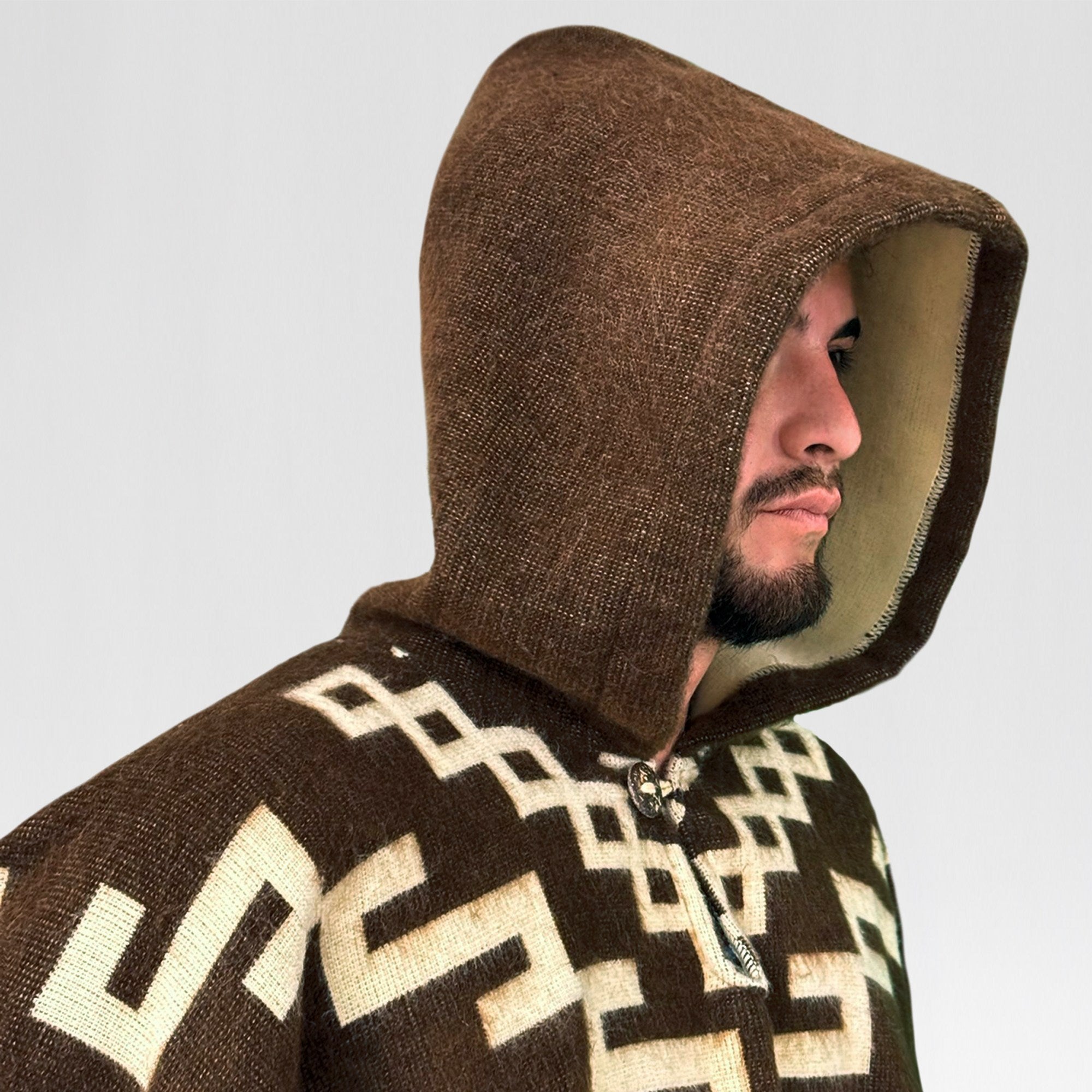 A person wears a brown hooded garment with geometric patterns, looking to the side. The background is plain and neutral.