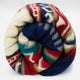 A colorful wool blanket is neatly rolled up, displaying a pattern of red, blue, green, and cream geometric shapes. It lies on a plain white background.
