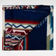 A folded blanket displays geometric patterns with zigzags and triangles in shades of blue, red, green, and white, set against a plain white background.