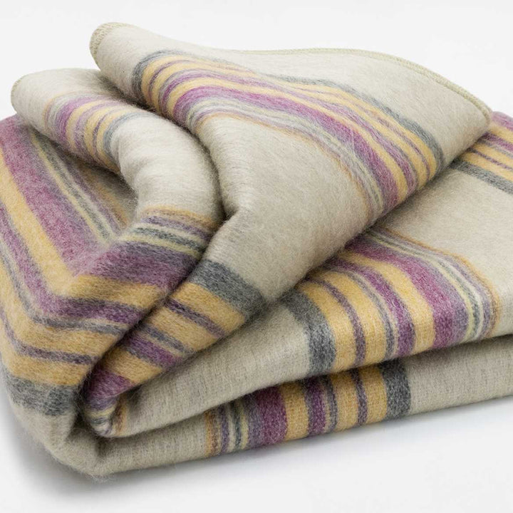 Quijos alpaca wool blanket – a folded woolen blanket displays yellow, purple, and grey stripes against a cream background, resting on a plain, neutral surface