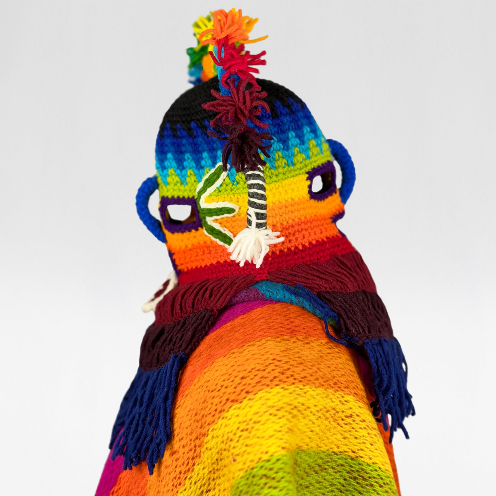 A colorful knitted figure displays vibrant rainbow-striped patterns. The figure features a hat with tassels on top and wrapped fabric resembling a scarf, against a plain white background.