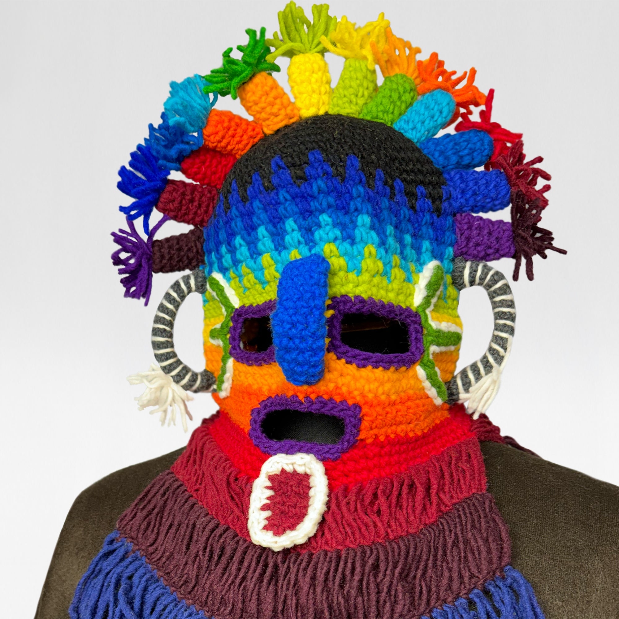 A multicolored, crocheted mask with a rainbow headdress and fringe accents is displayed against a neutral background. It features eye, nose, and mouth cutouts and striped circular side embellishments.