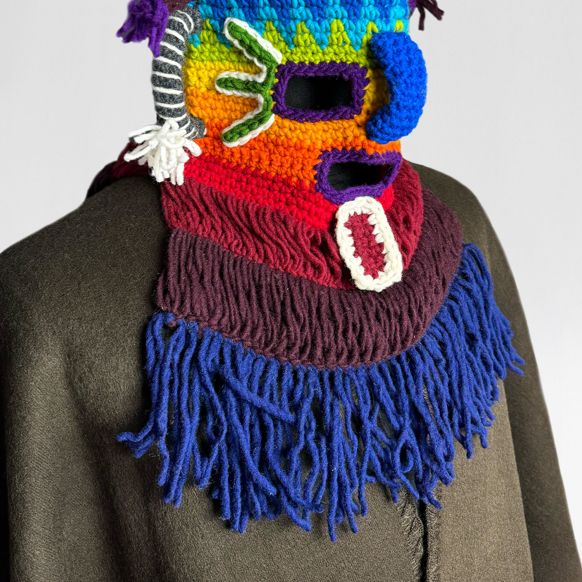 A multicolored knitted mask with eye and mouth openings features fringed edges and protruding shapes, worn over a dark coat in a neutral background.