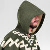 A person wears a green, patterned hooded garment, with their face partially obscured by the hood. The background is a plain light grey.