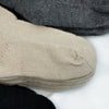 Socks are layered and overlapping, with a cream, dark grey, and black sock in a close-up view. The texture is knitted, and they are placed on a white surface.