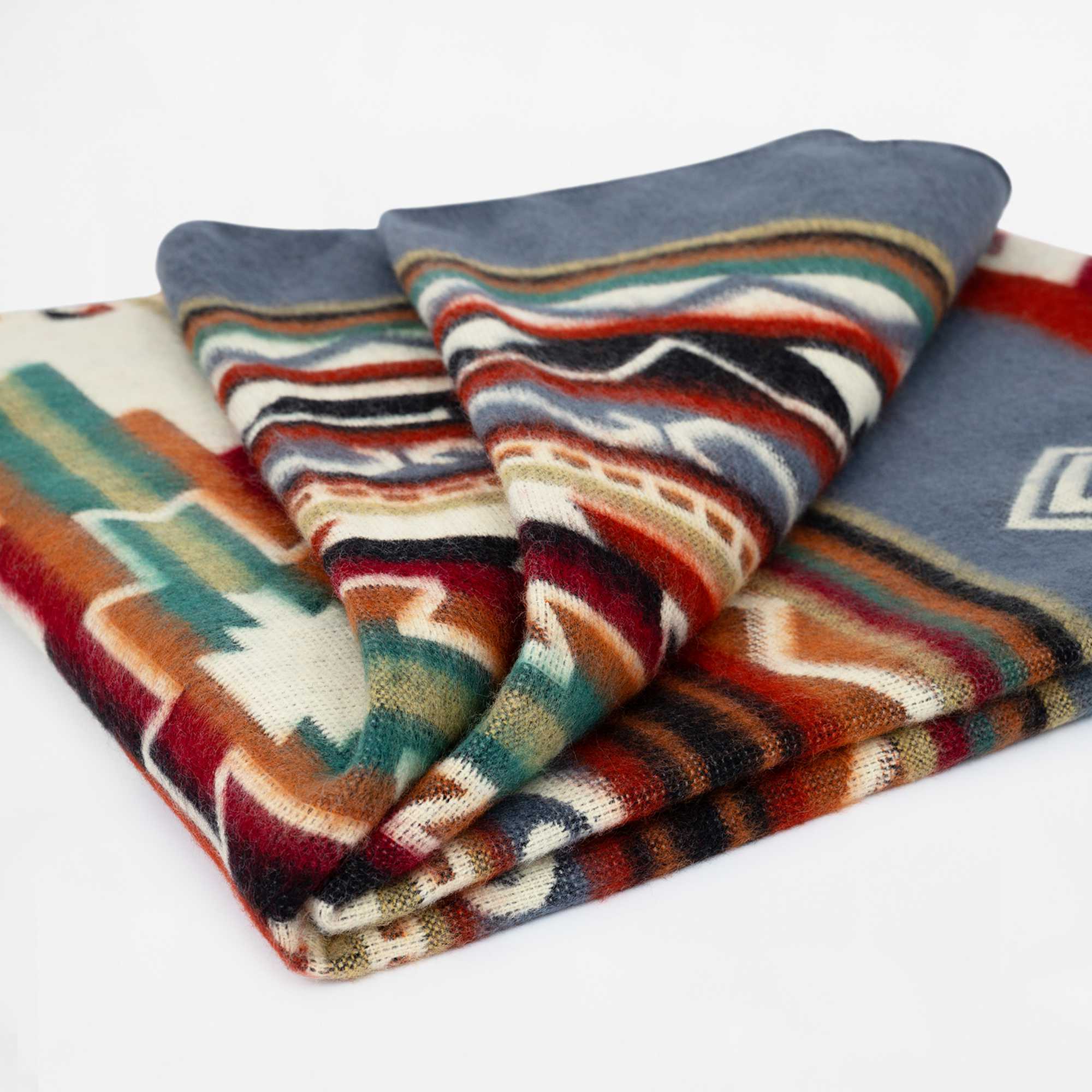 A folded woolen blanket displays vibrant geometric patterns in red, orange, teal, and cream, against a plain white background.