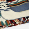 A blanket with a geometric pattern, featuring colors like red, green, and grey, is folded neatly. It rests on a white surface, showing detailed edging.