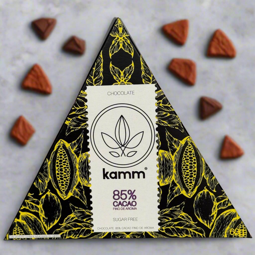 Triangular chocolate package labeled kamm, with 85% cacao, features intricate yellow floral designs. Surrounded by scattered triangular chocolate pieces on a gray background. Text: CHOCOLATE FINO DE AROMA SUGAR FREE.