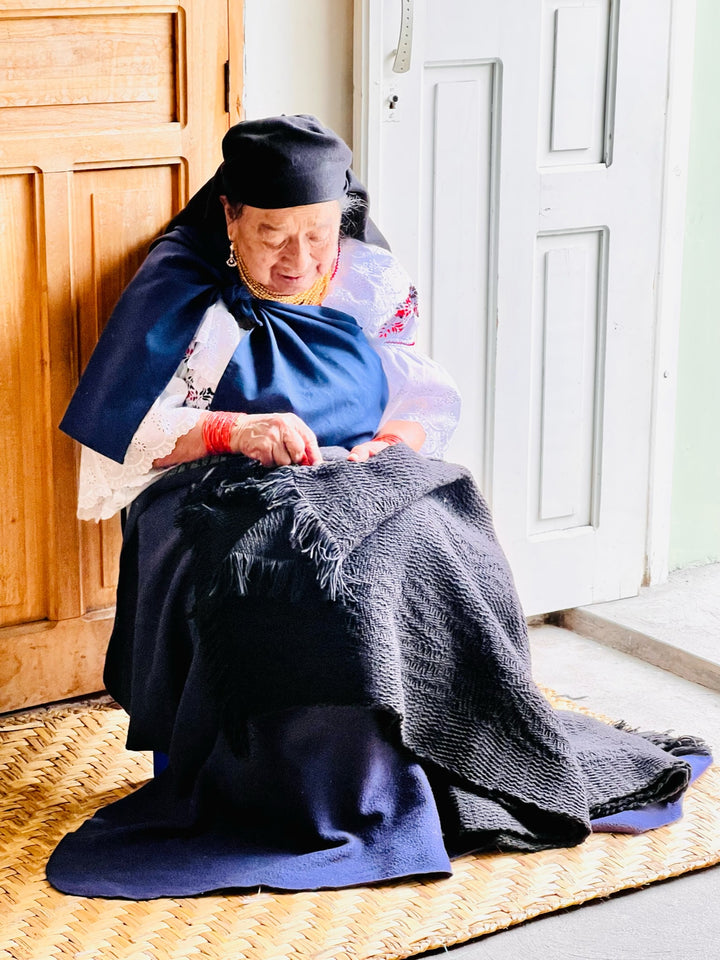 An elderly person sews a dark fabric while sitting on a woven mat by a wooden door inside a well-lit room. The person is dressed in traditional, colorful clothing.