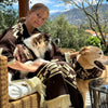 A person sits on a wicker chair, wearing a patterned brown poncho, hugging a small dog while another larger dog sits beside them. They are outdoors with a mountainous backdrop.