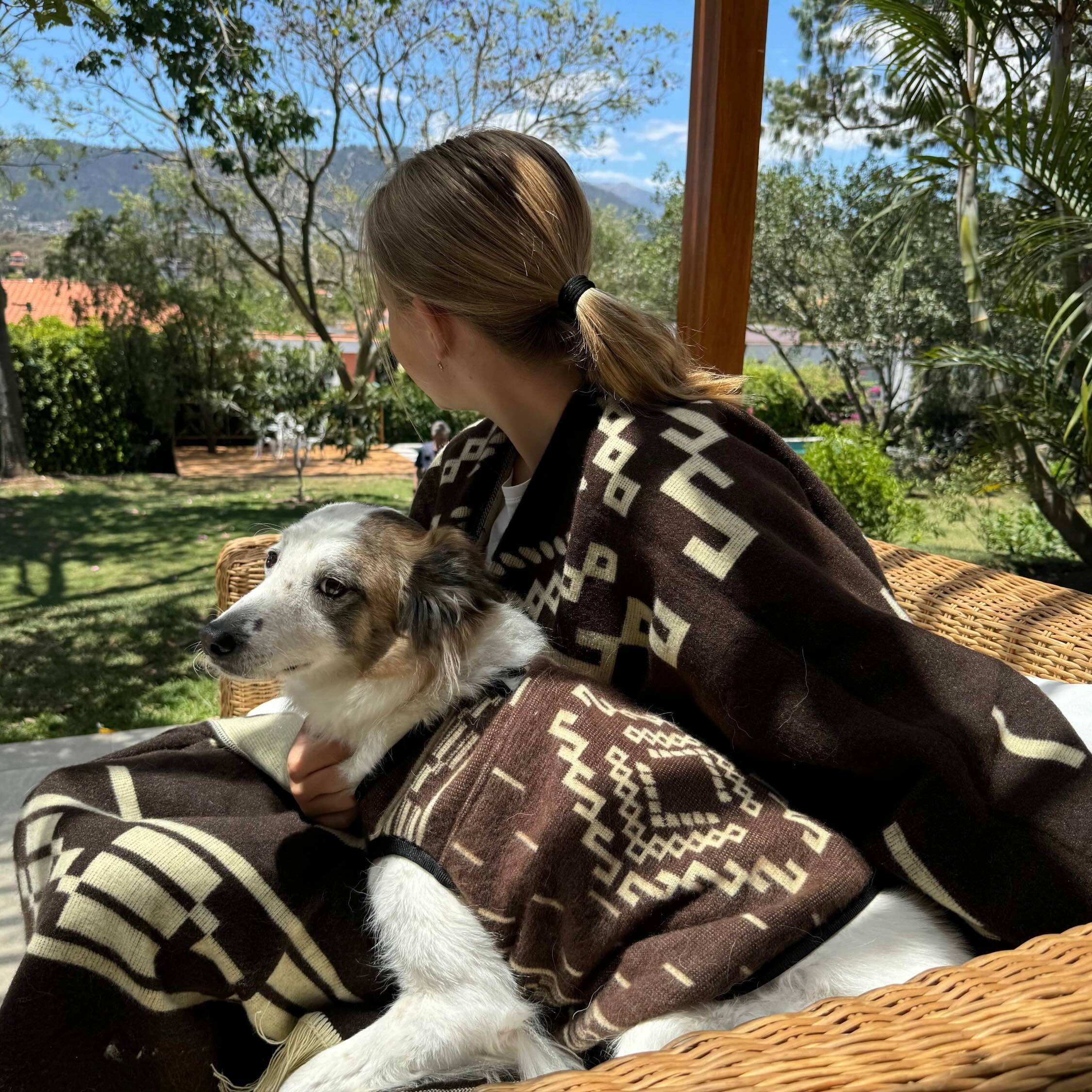 A person, wrapped in a patterned blanket, sits on a wicker chair holding a dog also in a similar blanket, looking towards a lush garden with trees and mountains.