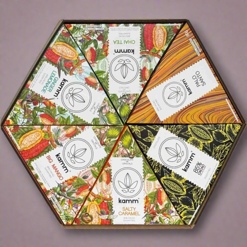 Hexagonal box containing six triangular chocolate sections, each uniquely wrapped: Chai Tea, Palo Santo, 85% Cacao, Salty Caramel, Big Mango, and Lemon Nuance. Surrounding design displays various colorful illustrations.