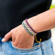 A multicolored woven bracelet rests on a person's wrist, as their hand is partially tucked into the pocket of blue jeans. The background is a blurred black fabric.