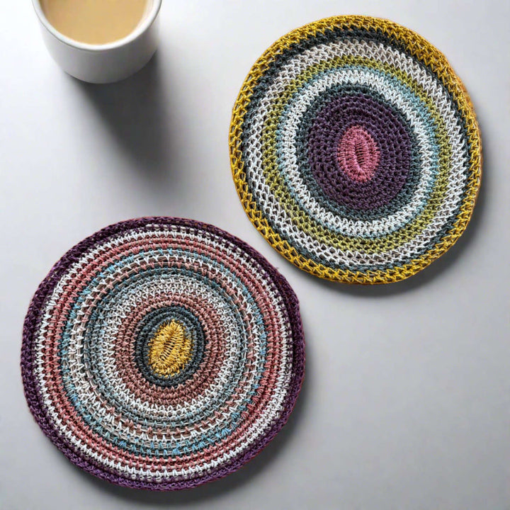 Two colorful, circular knitted coasters lie on a flat, neutral surface. A cup filled with a hot beverage is positioned nearby, adding a cozy, inviting touch to the scene.