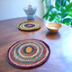 Two colorful, crocheted coasters rest on a wooden table, accompanied by a small wooden bowl and a glass vase. Green plants are visible in the softly lit background.