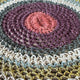 A circular, multi-colored woven rug displays concentric rings of pink, purple, green, cream, and yellow. It is lying flat on a wooden floor.