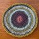A crocheted circular mat features concentric rings of pink, purple, green, and yellow hues, lying flat on a wooden surface.