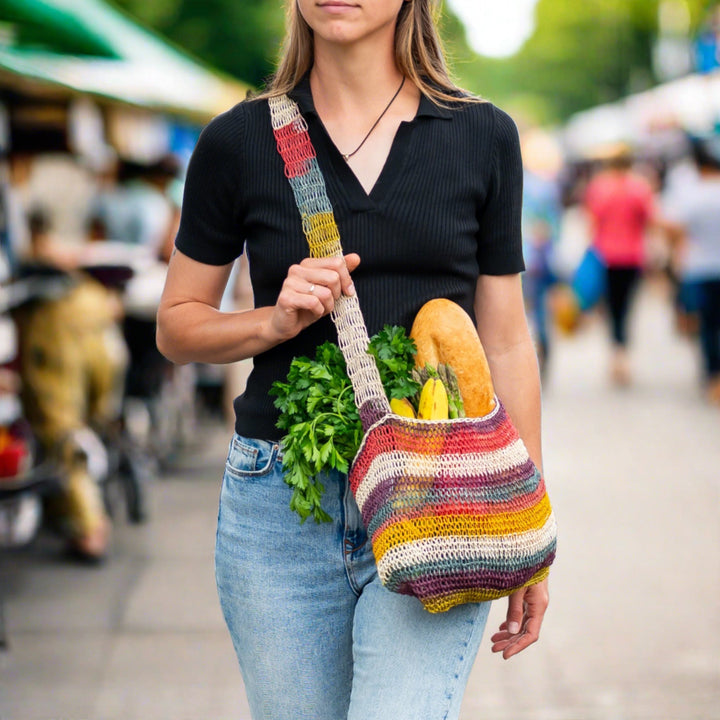 A person carries a colorful woven bag filled with groceries including bread, bananas, and leafy greens, walking through a bustling outdoor market with blurred shoppers in the background.