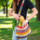 A person carries a colorful crocheted bag filled with groceries, including bread, herbs, bananas, and asparagus, while standing in a sunny park environment.