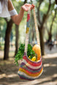 A multicolored woven bag, filled with fresh produce including bananas, bread, and leafy greens, is being held by a hand. The background shows a sunlit, tree-lined path.
