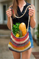 A colorful, knitted bag carries a baguette, asparagus, bananas, and parsley. A person holds it with both hands on a street, creating a casual shopping scene.