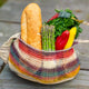 A woven, multicolored bag holds a baguette, asparagus, leafy greens, and assorted peppers, resting on a wooden surface outdoors.