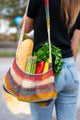 A multicolored net bag holds a baguette, asparagus, bell peppers, and parsley as a person walks on the street, wearing jeans and a black top.