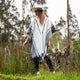 A person wearing a hat and patterned poncho walks through tall grass in a lush, wooded area, surrounded by trees and vegetation.