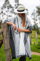 A person in a white hat and patterned gray poncho leans against a wooden fence, holding sunglasses, with a blurred green outdoor landscape in the background.