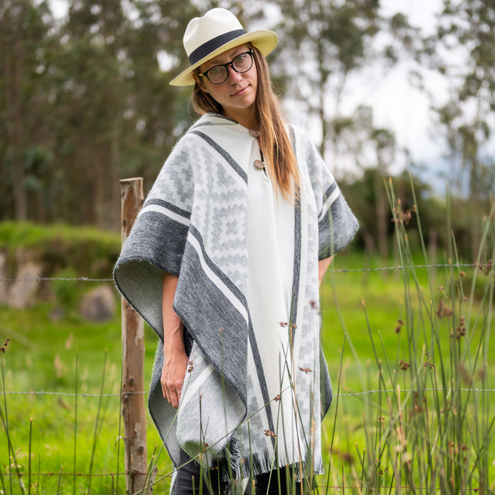 A person in glasses and a white hat is wearing a gray and white poncho. They're standing in a grassy field with trees and a wooden fence in the background.