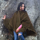 A person wearing a dark green poncho stands against a large tree trunk, with one hand touching the tree. The individual is dressed in jeans and a pink top.