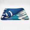 A folded blanket displays a geometric pattern with shades of blue, teal, and gray, featuring white accents. It's positioned against a neutral background, emphasizing its vibrant design.