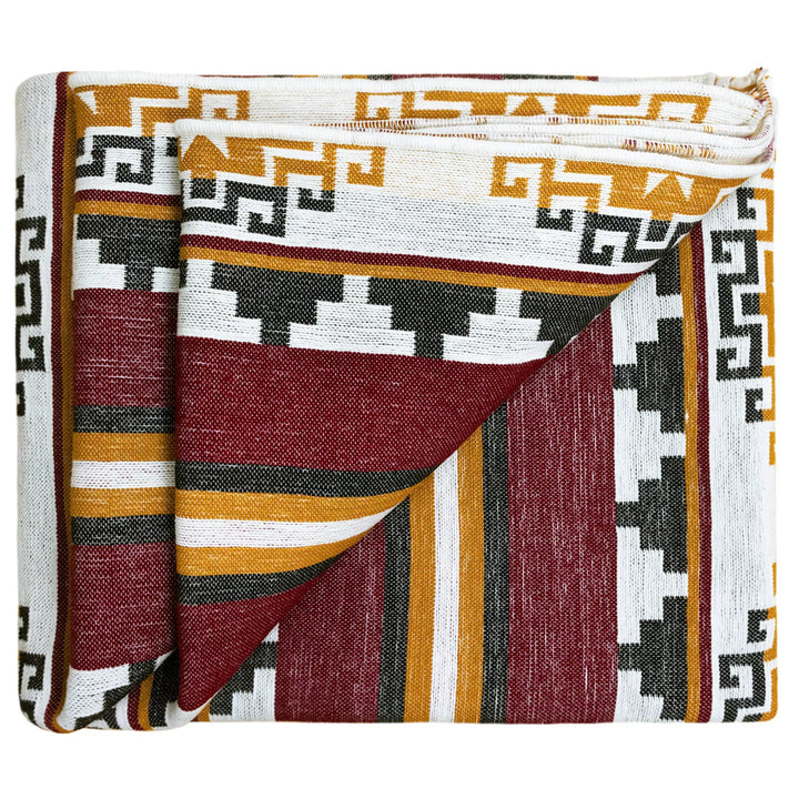 A folded textile with geometric patterns, featuring red, yellow, white, and black colors, lies neatly, emphasizing intricate, symmetrical designs in an abstract, cultural motif on a plain background.