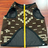 A patterned brown vest lies flat on a wooden floor. A yellow measuring tape runs vertically along its center, indicating measurements. The vest features geometric designs in beige with quilted black lining.