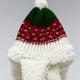 A knitted hat with green, red, and white patterns, topped by a white pom-pom, rests on a fluffy white material, set against a plain background.
