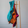 Colorful, patterned blankets drape over a wooden rack. The blankets are vibrant shades of turquoise, orange, and magenta, displaying geometric designs. The setting is minimal with a plain background.