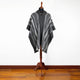 A gray and white striped poncho, draped over a mannequin, is displayed on a wooden stand against a plain white background with a wooden floor.