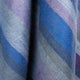 Morona baby alpaca wool blanket – striped fabric drapes vertically, featuring alternating soft hues of blue, purple, and gray. The fibers are visibly textured, indicating a cozy, wool-like material in gentle folds