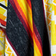 A woven fabric drapes, showcasing bold stripes of black, yellow, orange, and red, with intricate geometric patterns. The context suggests a vibrant textile design.
