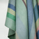 Sucua baby alpaca wool blanket – a soft, draped fabric displays pastel green, blue, yellow, and pink diagonal stripes. The weave and texture suggest warmth and comfort. The background is a plain, light color