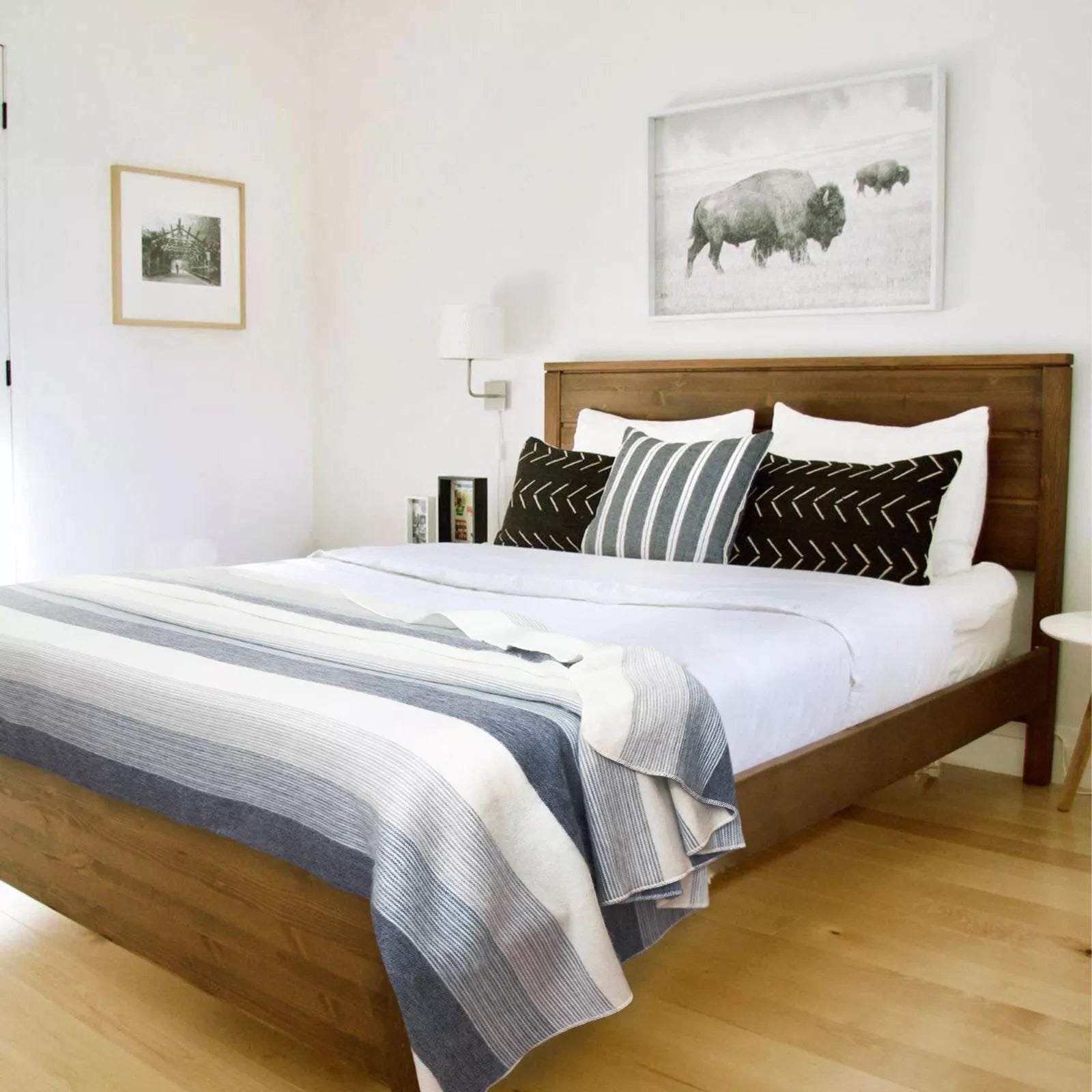 Wooden bed neatly arranged with pillows and a striped blanket; set in a bright room with hardwood floors, wall art of bison, and a small wall lamp nearby.