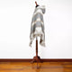 A hooded, fringed poncho drapes over a wooden mannequin stand, displayed against a plain white wall on a polished wooden floor.