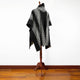 A black poncho with white geometric patterns hangs on a wooden stand in a room with a wooden floor and plain white wall.