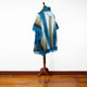 A fringed, multicolored poncho drapes over a wooden mannequin, standing on a polished hardwood floor against a plain white wall.