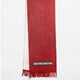A red scarf with fringe hangs vertically, featuring a small black and white geometric pattern near the bottom. It contrasts against a plain white background.