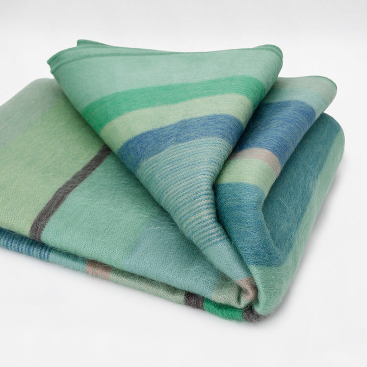Sucua baby alpaca wool blanket – a folded, soft blanket displays green, blue, and gray horizontal stripes. It rests on a plain, light background, emphasizing its cozy and inviting texture