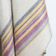 Quijos alpaca wool blanket – a woolen blanket displays horizontal stripes in purple, yellow, and gray, draped in overlapping layers against a plain backdrop