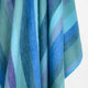 Pululahua baby alpaca wool blanket – a piece of fabric hangs with broad, vertical stripes in various shades of blue, teal, and purple, providing a soft, soothing texture against a plain background